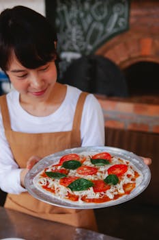 Asian female chef showcases a freshly prepared pizza in a rustic restaurant kitchen setting.