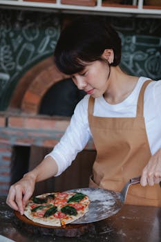 Asian woman in apron preparing delicious homemade pizza with fresh ingredients in a traditional kitchen setting.