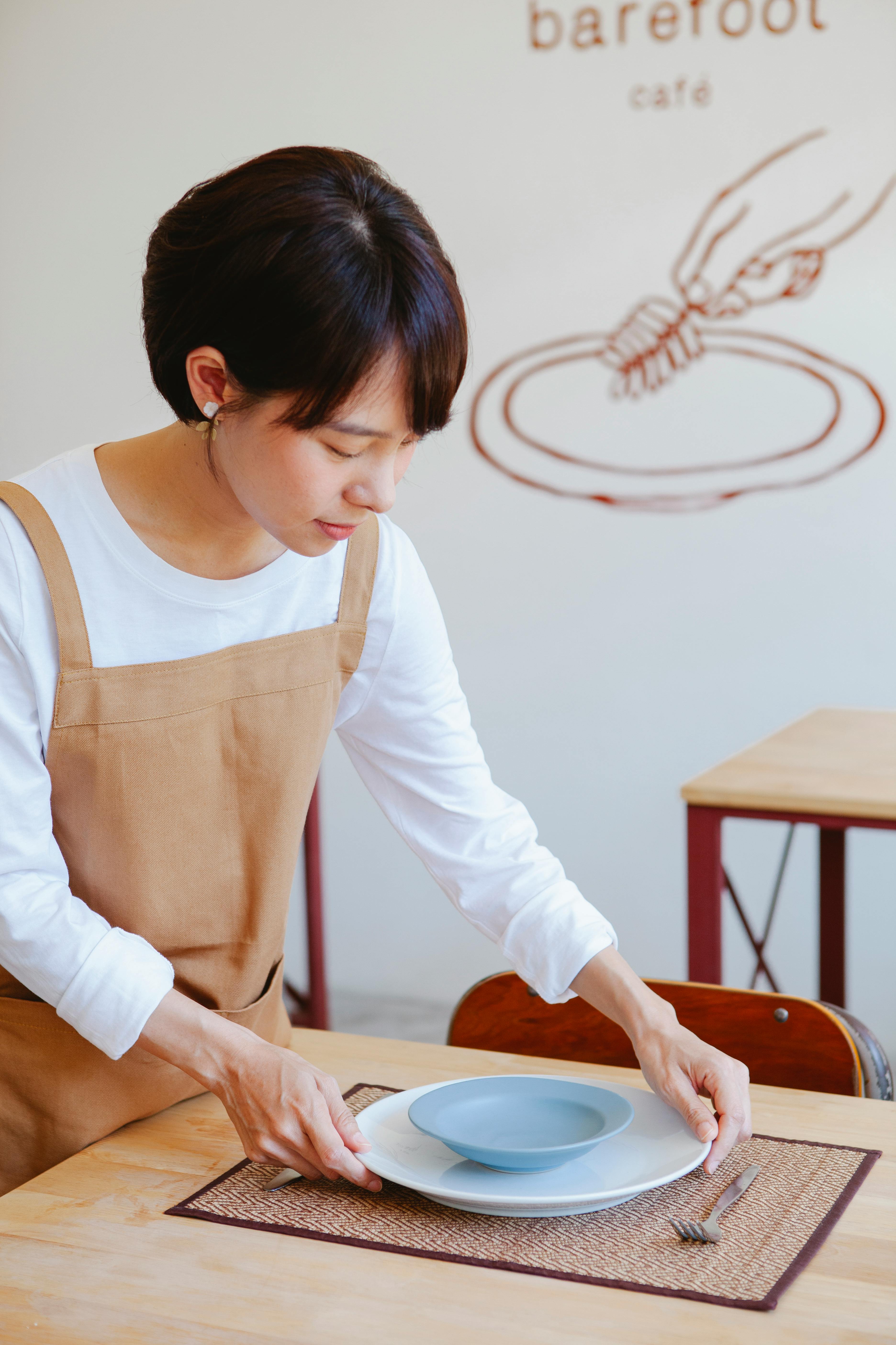 Asian woman arranging a table setting at a minimalist cafe, enhancing ambience.