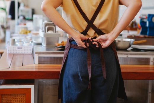 Person seen from behind tying a denim apron with brown crossed straps