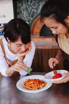 Two women creatively garnishing and plating a pasta dish in a kitchen setting.