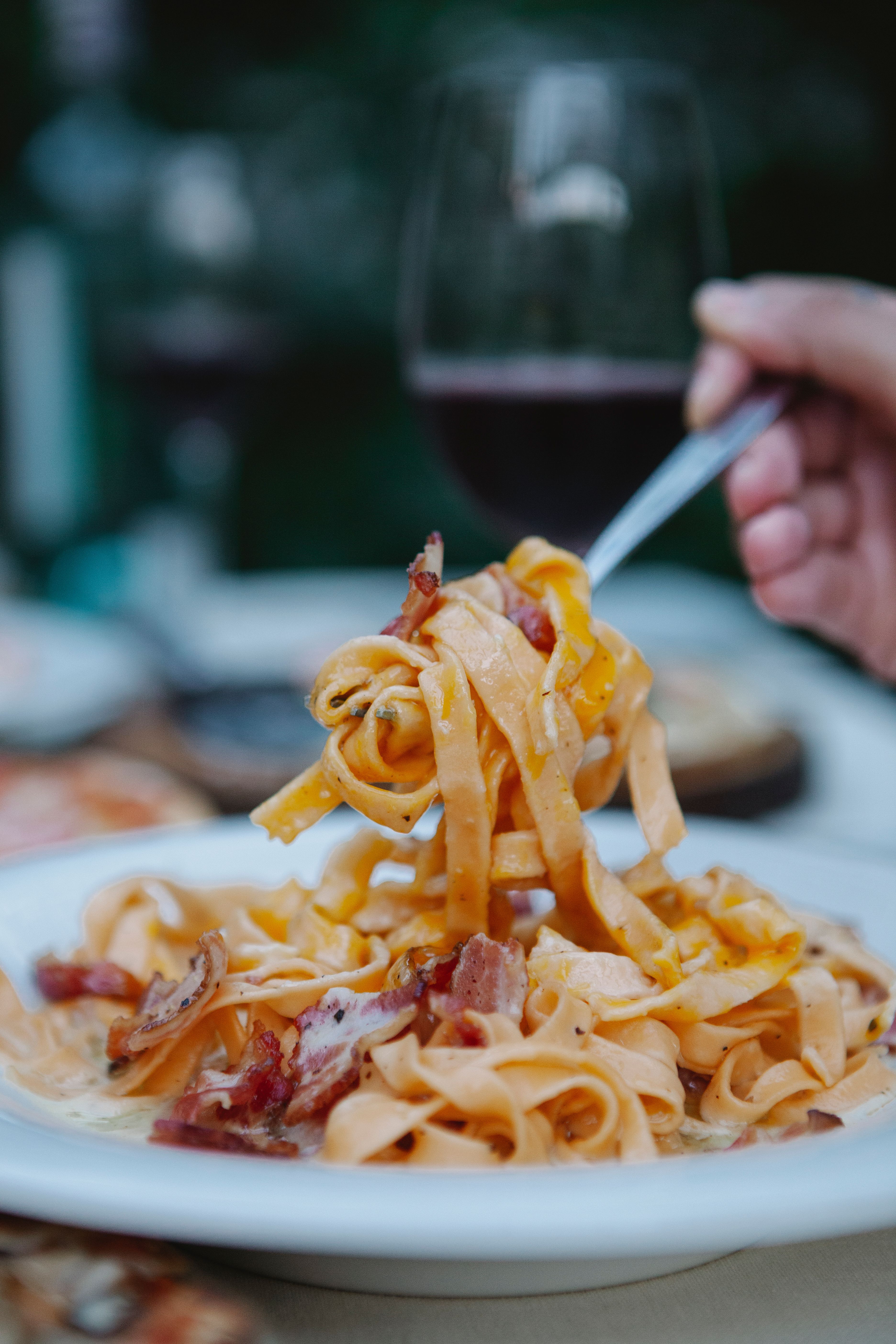 A tantalizing close-up of pasta and bacon on a fork with red wine in the background.