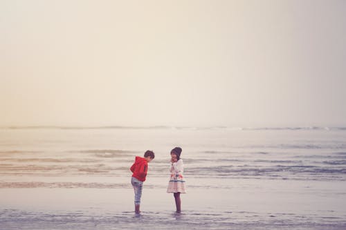 Side view of unrecognizable little ethnic boy and girl in stylish outfits standing in water of waving ocean against cloudy sunset sky