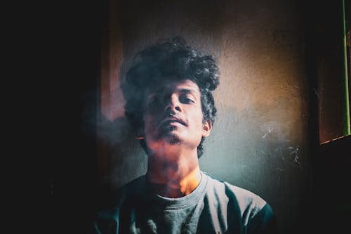 Confident male with curly hair looking at camera while standing near shabby wall in dark room