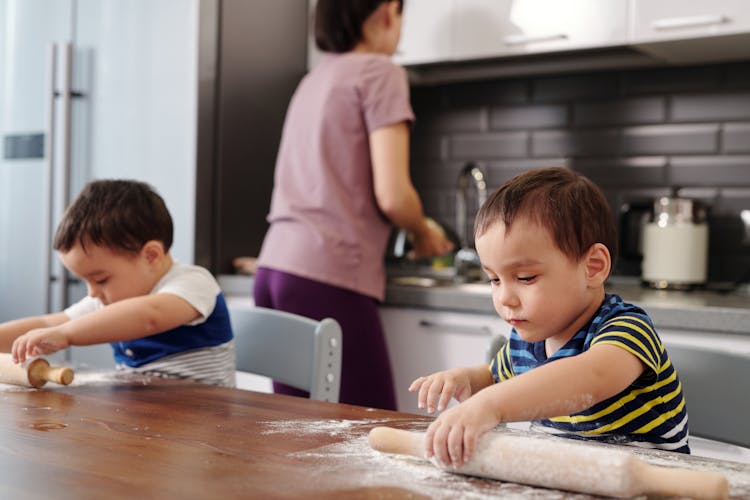 Two Little Boys Helping Their Mother In The Kitchen And Using A Rolling Pin 