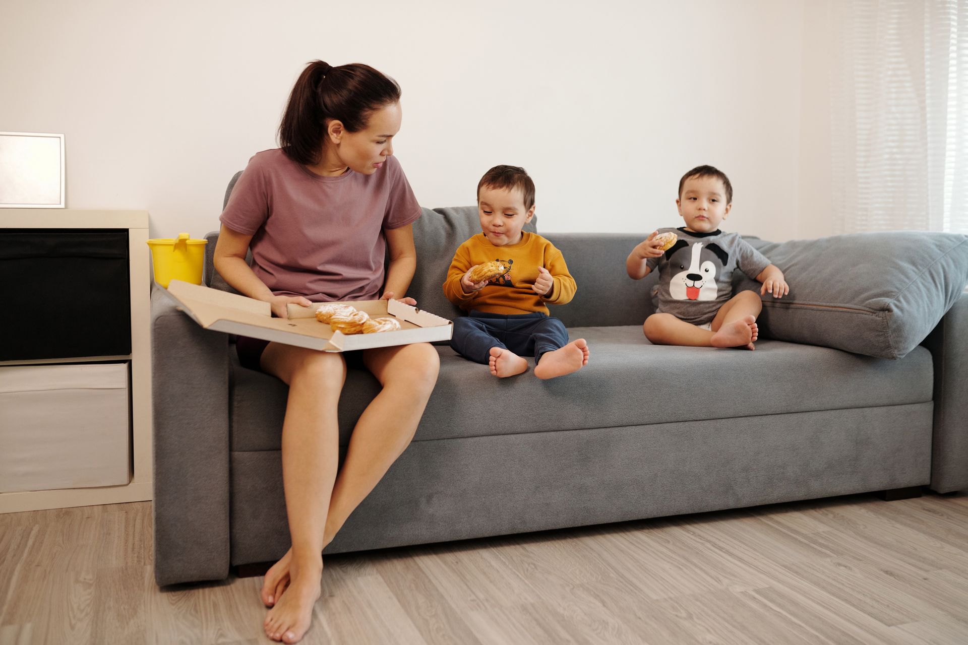 A mother and her twin toddlers enjoying croissants on a sofa, capturing a cozy family moment.