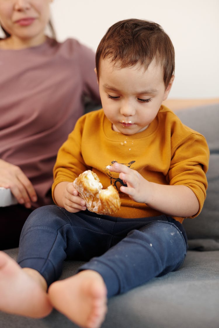 A Little Boy In Brown Long Sleeves Eating Bread While Sitting On A Couch