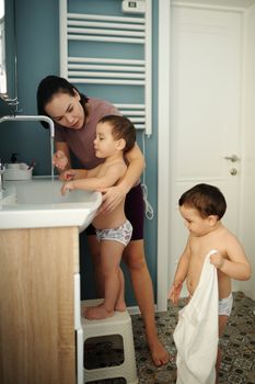 A mother assists her young sons with washing hands at a sink in a home bathroom.