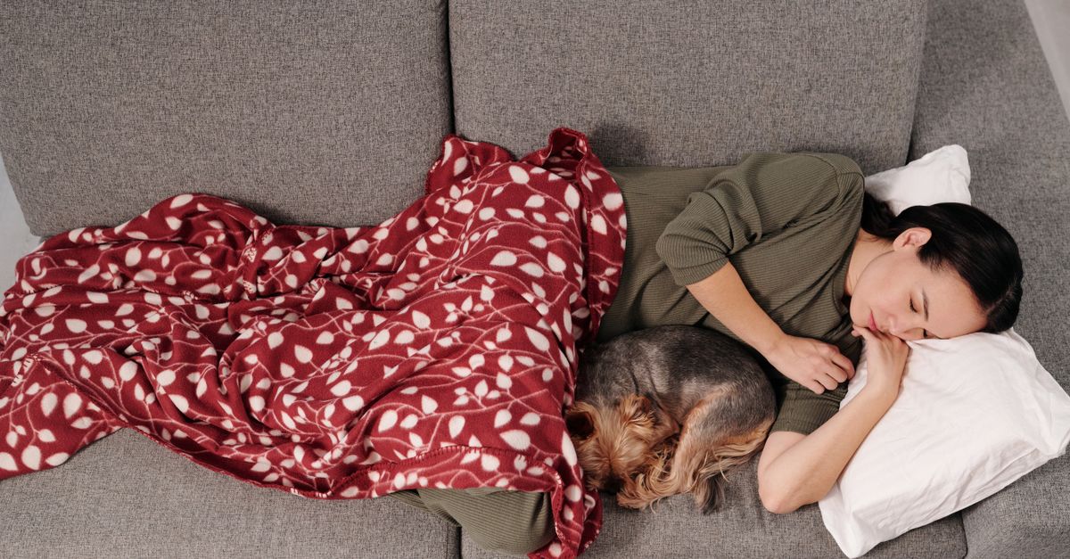 Uma mulher e seu cachorro dormindo pacificamente em um sofá cinza com uma manta vermelha.
