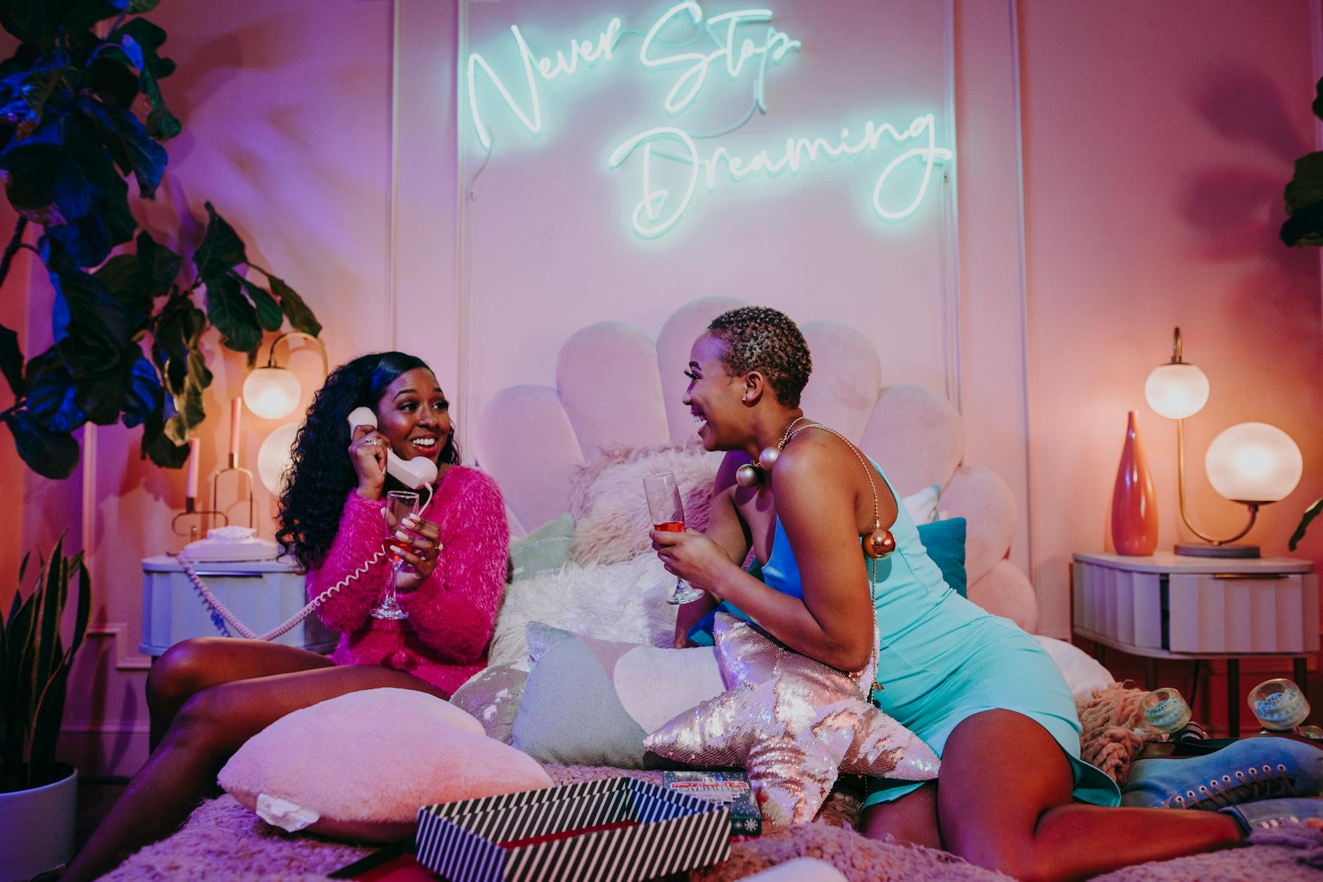 Two women enjoying a cozy indoor pajama party with a neon 'Never Stop Dreaming' sign.