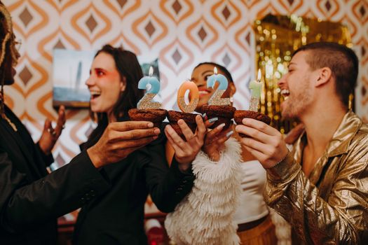 Group of friends joyfully celebrating New Year's Eve with cupcakes and candles indoors.