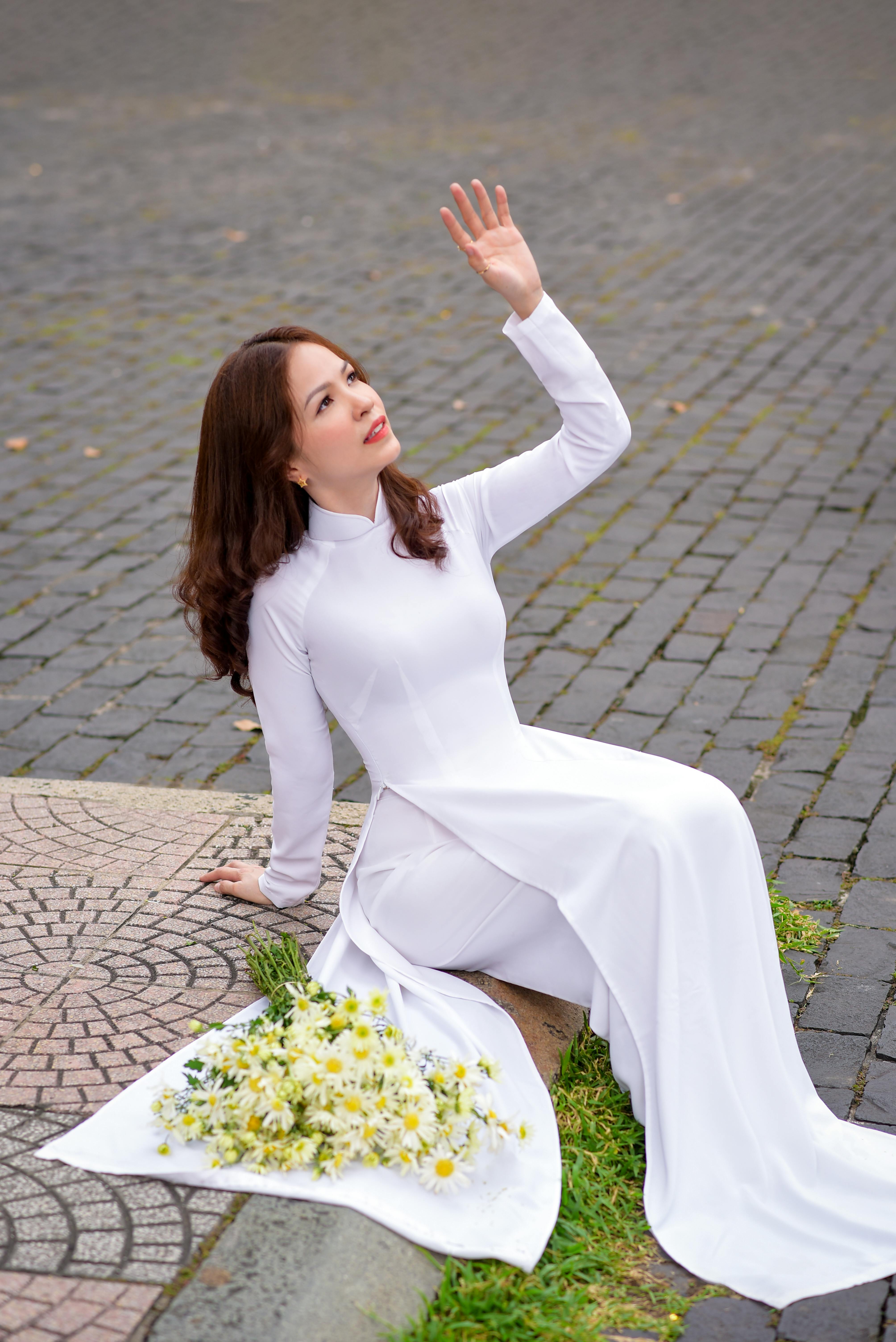 a woman wearing white ao dai sitting beside bunch of white flowers while looking afar