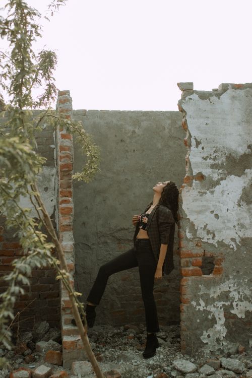 Free Full body side view of young lady in trendy outfit standing near shabby walls of broken abandoned building near green plant in daytime Stock Photo