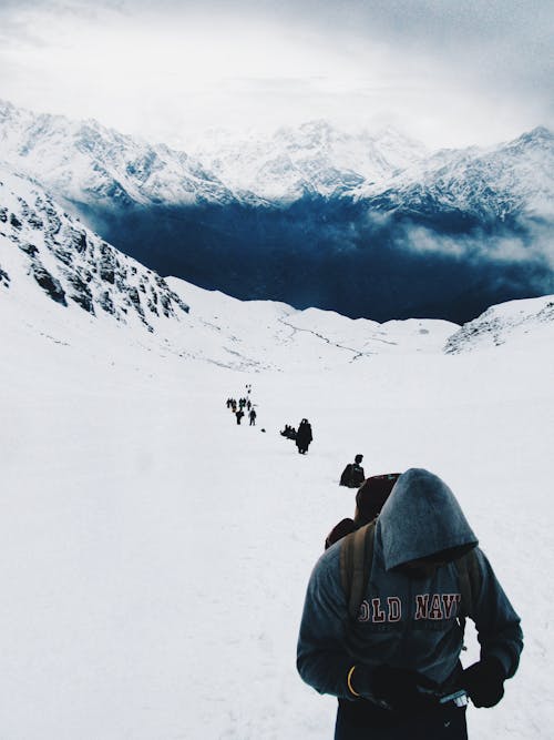 Vertical Shot of a Man in a Hood and People Walking behind in Snowed Mountains with Blue Lake