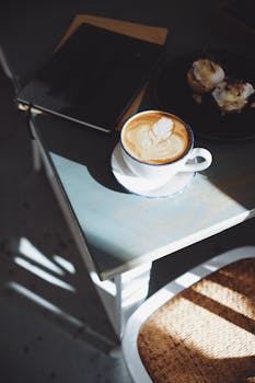 A cup of latte with beautiful art beside pastries on a sunlit table, evoking a cozy coffee shop vibe.