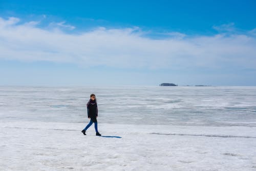 Side view full body of anonymous female in warm outfit strolling on icy shore near endless frozen sea under blue sky in daylight