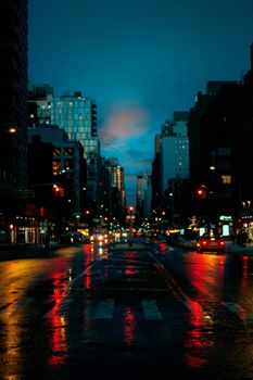 Asphalt road with crosswalk and glowing vehicles near buildings in twilight under blue cloudy sky in city district