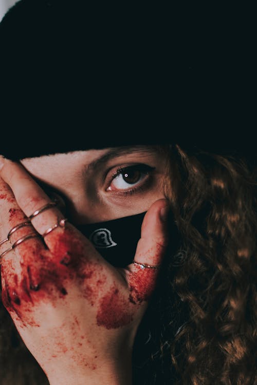Free Unrecognizable female in black mask and hat covering face with hand in blood and looking at camera Stock Photo