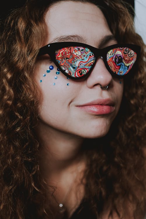 Closeup of cheerful young female in sunglasses with vivid multicolored lenses with ring in nose and rhinestones on face looking away