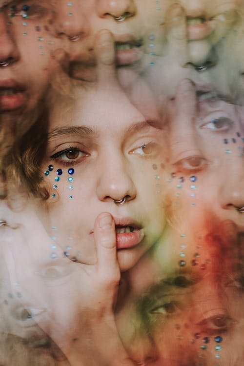 Abstract kaleidoscopic portrait of young lady with rhinestones on face and piercing in nose with makeup touching lips with finger and looking away thoughtfully