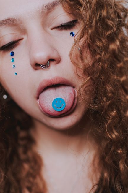 Crop female with piercing in nose and blue smiley sticker on tongue and rhinestones on cheeks with closed eyes in light place