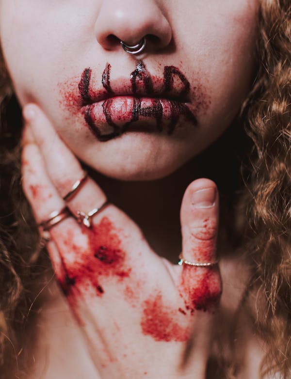 Crop anonymous female victim wearing rings and piercing in nose while touching cheek with hand with blood and wounds on knuckles and word liar written with marker on bloody lips