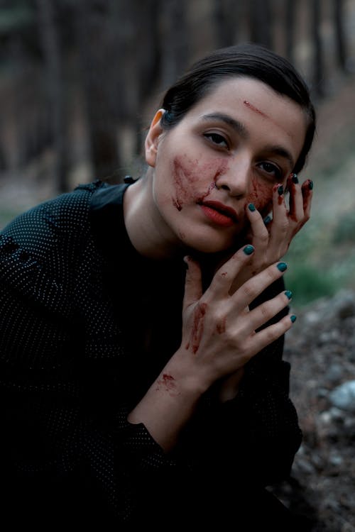 Calm attractive female in black clothes touching face covered with dirt and looking at camera while sitting on ground in woodland