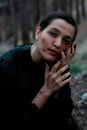 Calm attractive female in black clothes touching face covered with dirt and looking at camera while sitting on ground in woodland