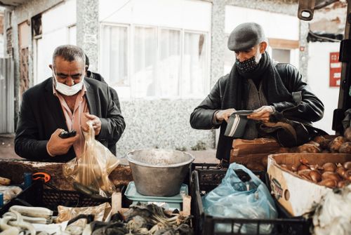 Free Multiracial male in outerwear and protective masks standing near stall with different products in boxes selling food to buyer in local street market Stock Photo