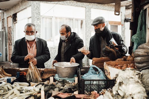 Free Multiethnic male buying food from seller in outerwear and protective mask near stall with different vegetables in boxes near metal basin and packets in local market in city street Stock Photo