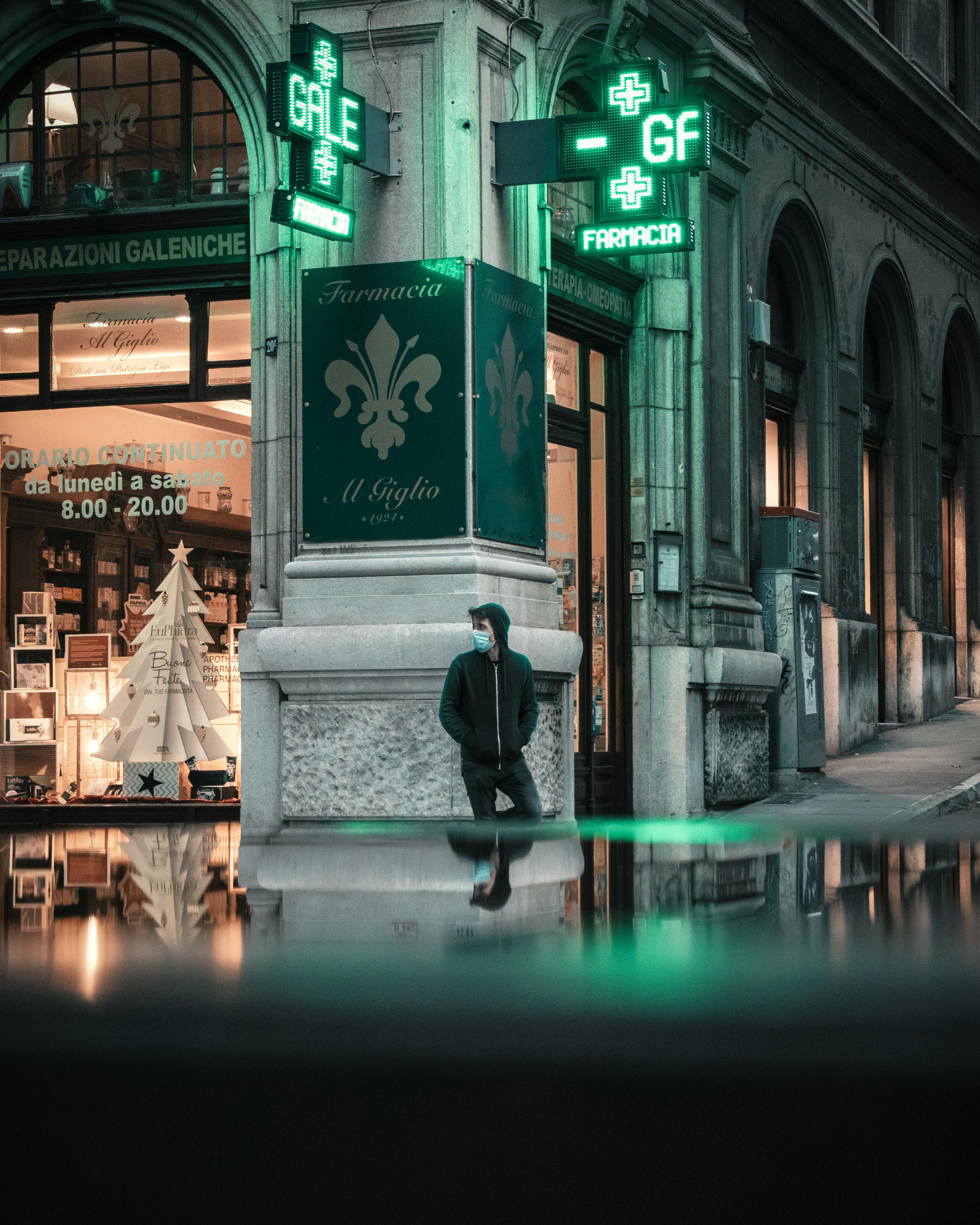 A person stands by a neon-lit pharmacy at night, reflecting urban city's vibrant life.