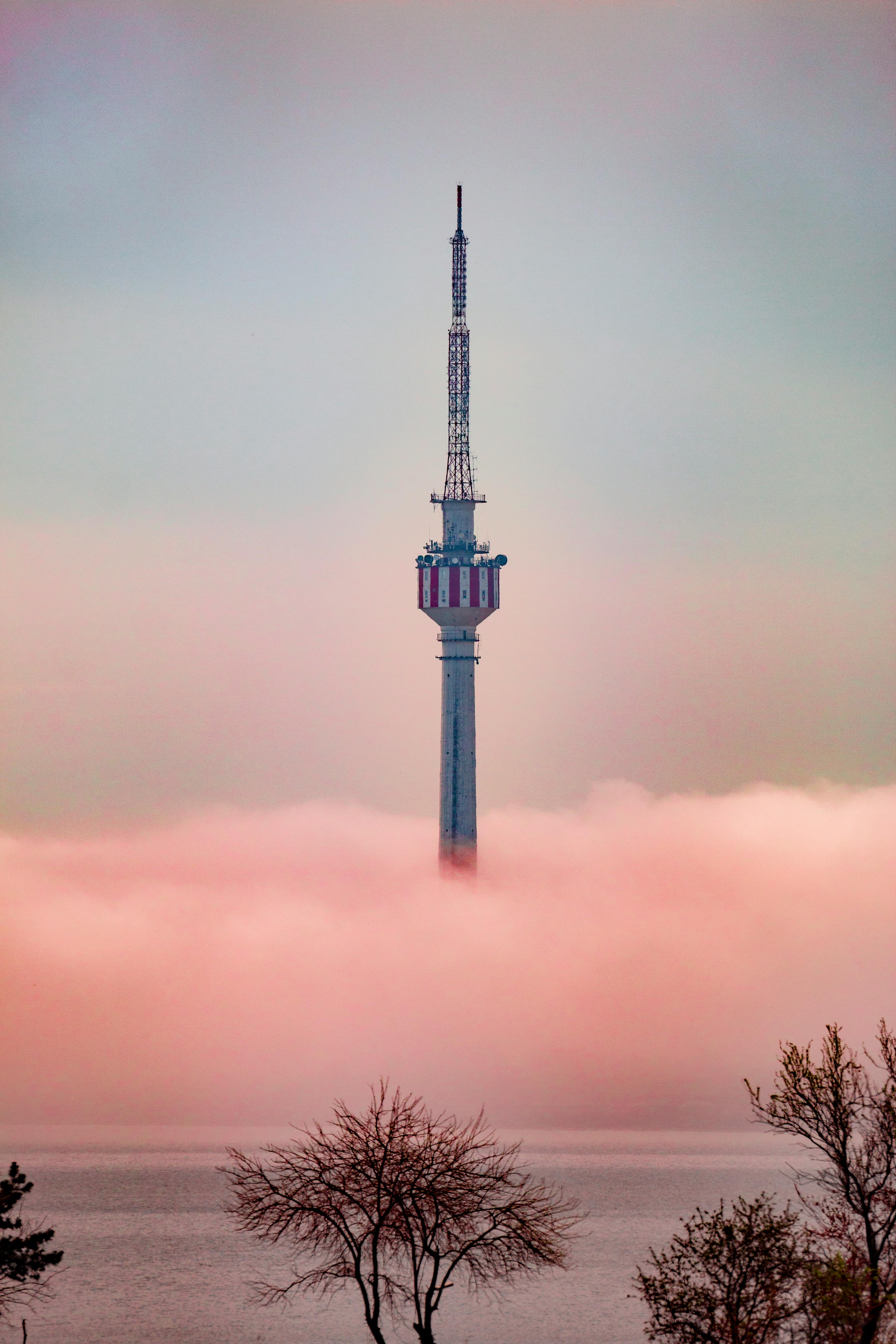 A TV tower emerges through a dense fog over a serene lake, surrounded by trees.