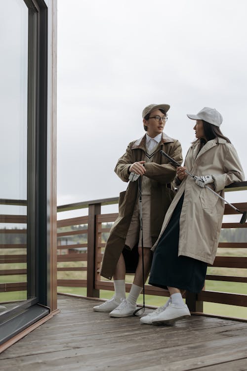 Man and Woman in Beige Coats talking on a Terrace while Holding Golf Clubs 