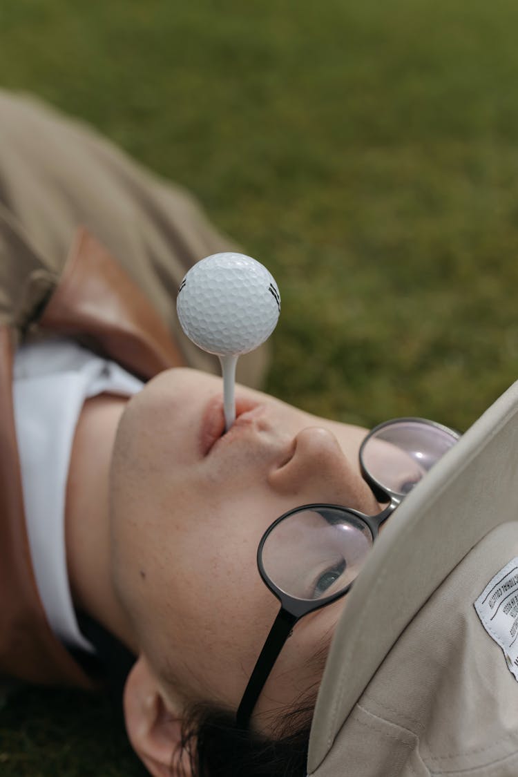 Person Lying Down On A Grass Holding Golf Ball On His Lip