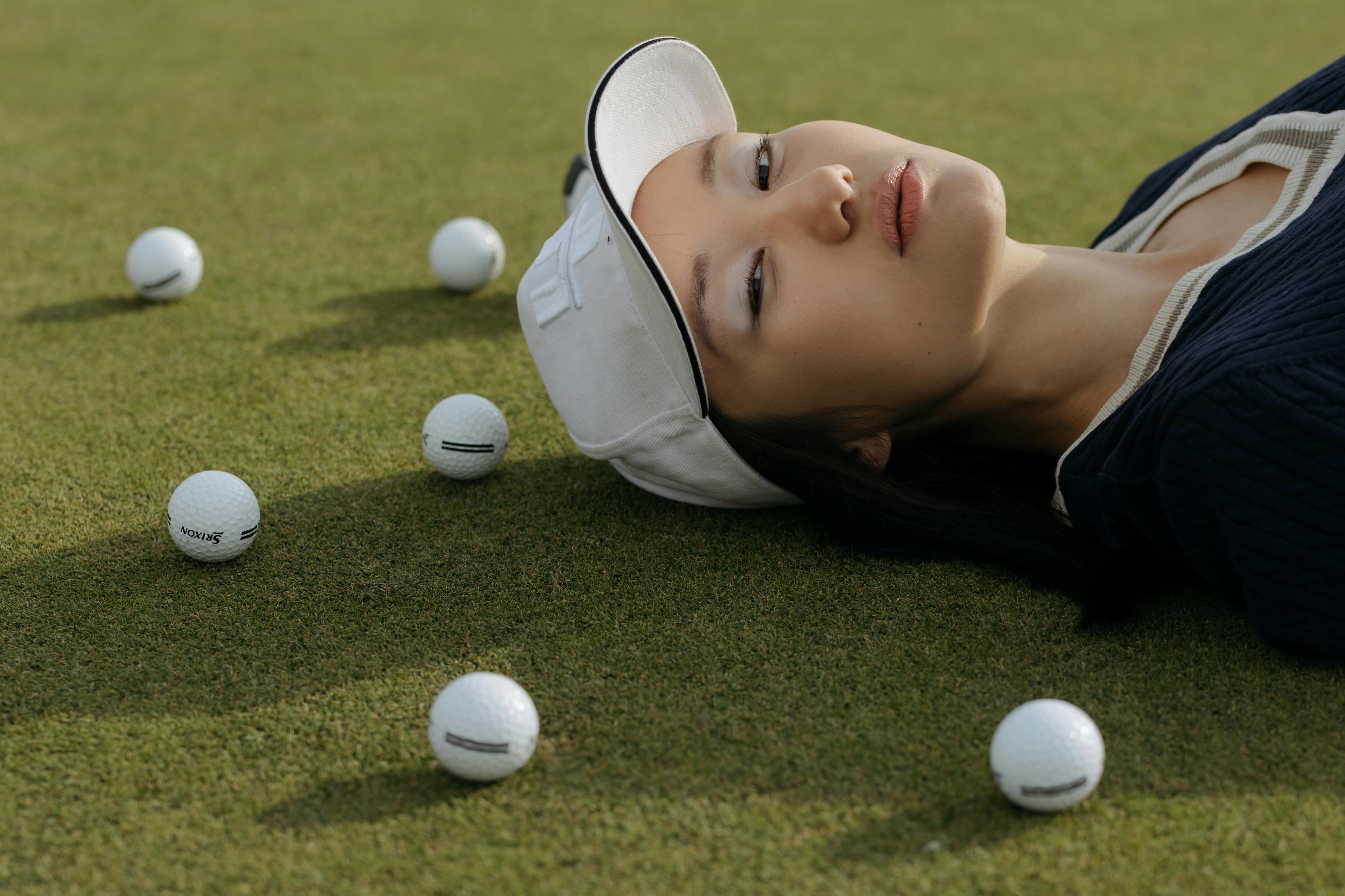 A woman in a white cap lying on grass with scattered golf balls, embodying leisure and style.