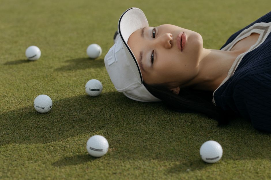 A woman in a white cap lying on grass with scattered golf balls, embodying leisure and style.
