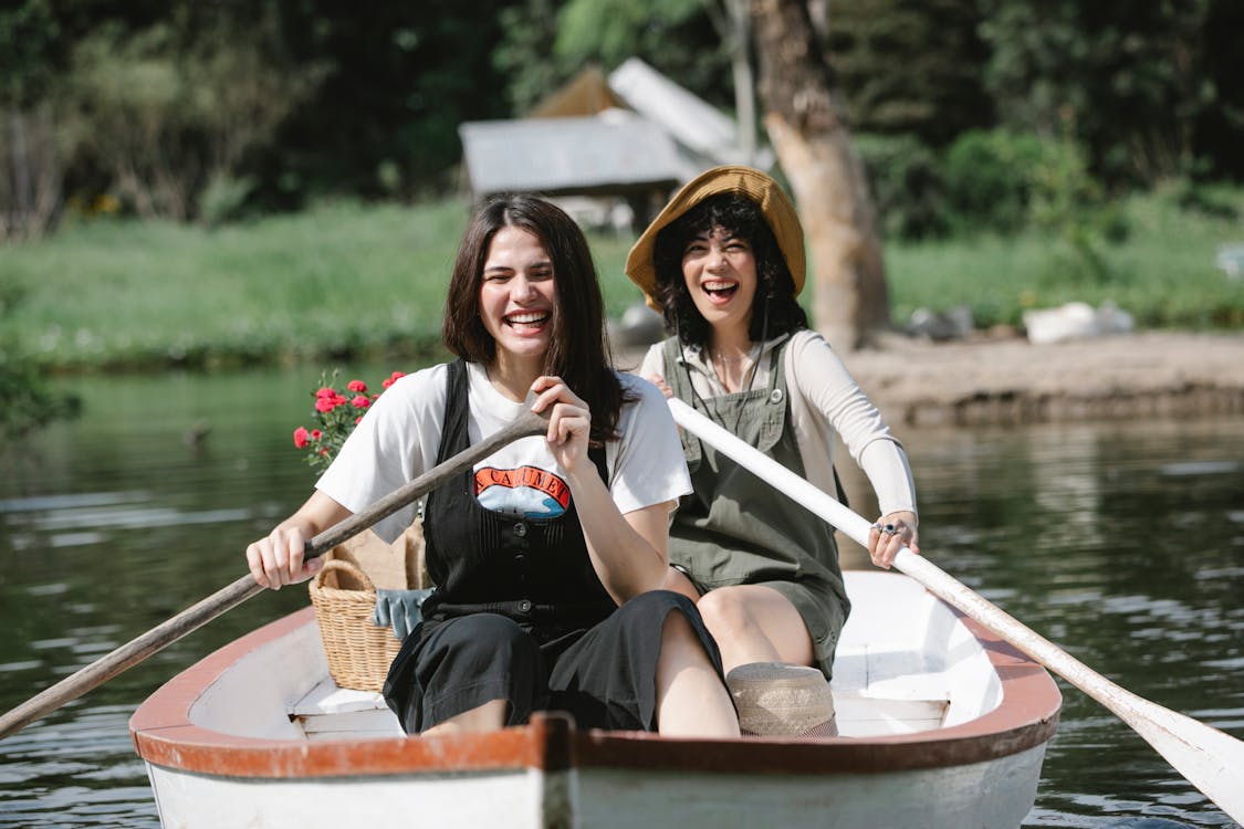 Free Happy young female friends in overalls sitting in boat near basket with flowers and using paddles while floating in pond near green grassy coast with trees in summer sunny day Stock Photo