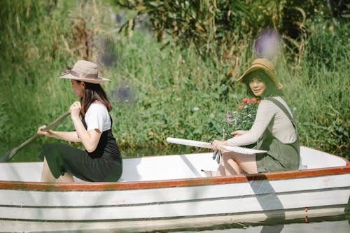 Free Side view of young women in overalls and hats sitting in boat while floating in lake near flowers and using paddles near grassy shore with bushes in sunny summer day Stock Photo