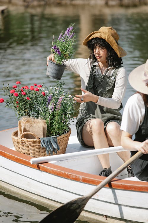 Young women gardeners in overalls and hats in boat near basket with potted flowers and gloves using paddles while floating on lake in summer sunny day in countryside