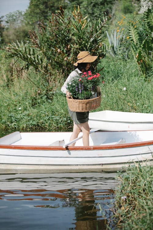Side view of anonymous female gardener in overall and hat with wicker basket with flowers in boat near riverside with green grass and plants in sunny summer day in countryside