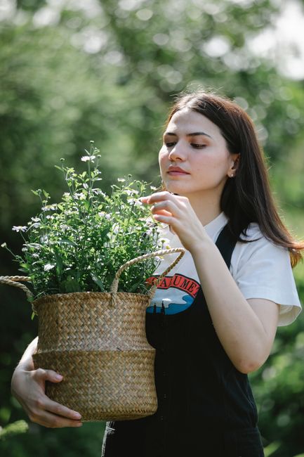 Young female in casual clothes standing with wicker basket with flowers in garden in sunny summer day