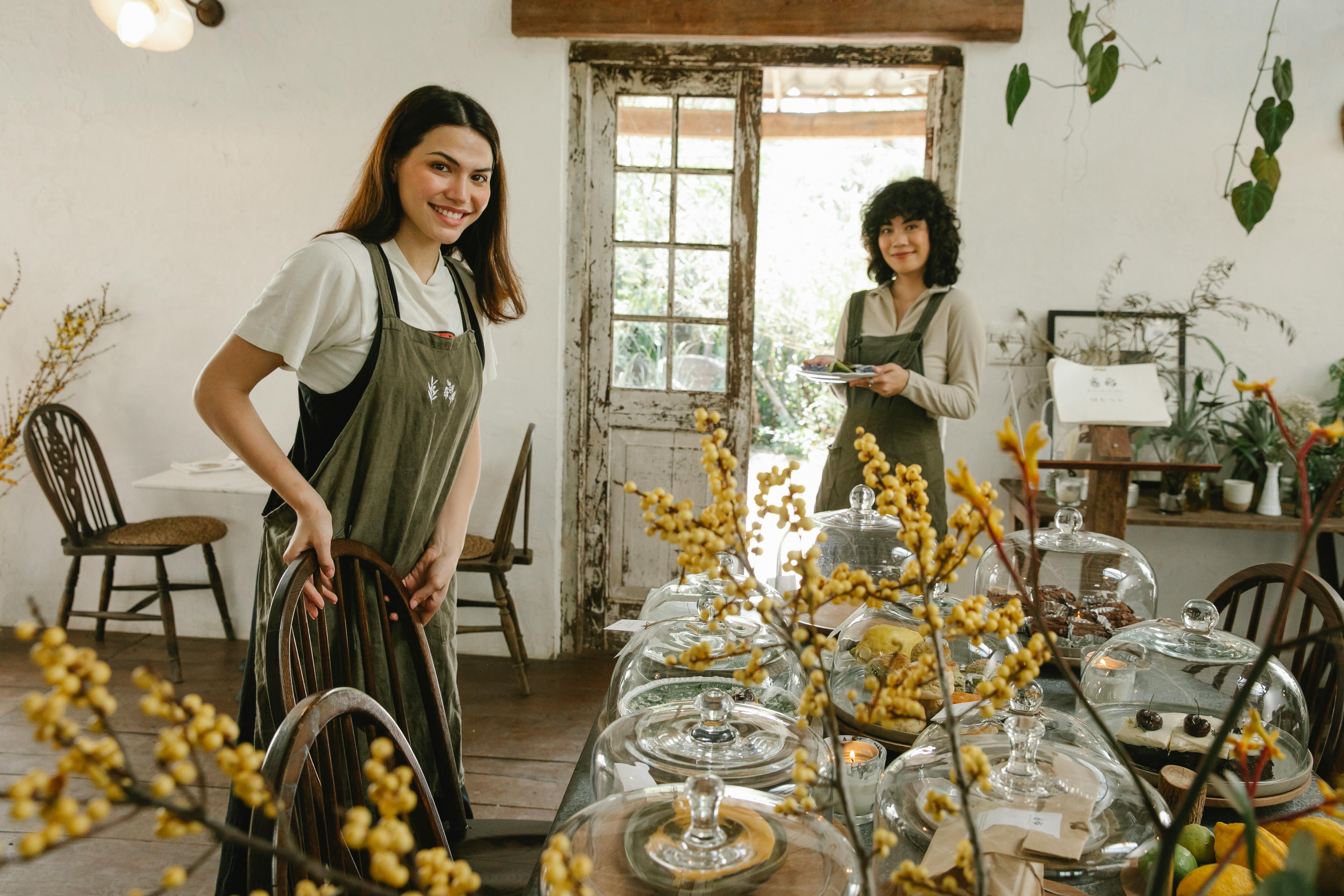 Positive waitress in aprons serving table with various sweet cakes in cozy cafeteria with light walls and plants and looking at camera