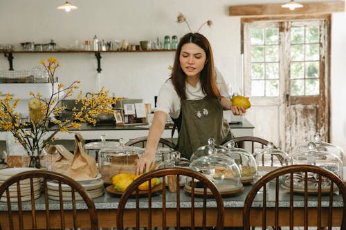 Free Concentrated waitress in apron standing and serving table with various delicious cakes in spacious cafe in daytime Stock Photo