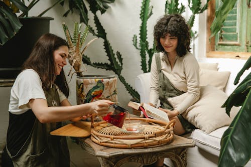 Smiling waitress in apron pouring hot tea into cup for female client sitting with book on couch in cafe