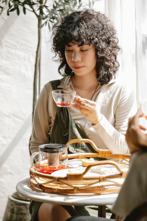 Free Curly haired female with glass cup of herbal tea sitting at table with friend Stock Photo