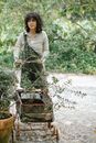 Ethnic female gardener rolling wooden cart in garden with plants in clay pots