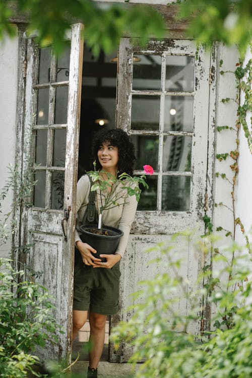 Positive ethnic female gardener standing with blooming potted plant in weathered entrance of rural house