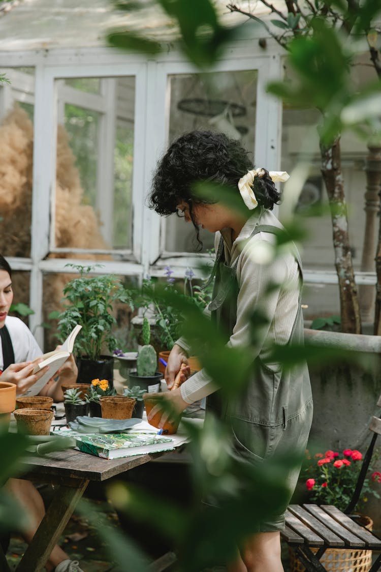 Female Gardener Planting Flower In Pot With Colleague Reading Book