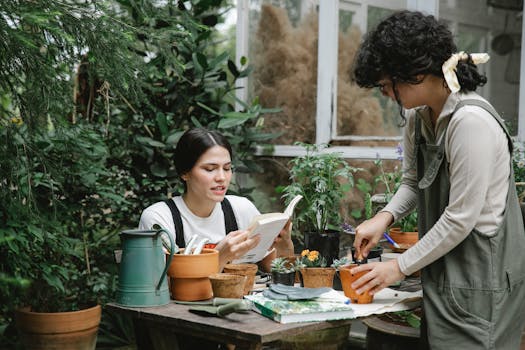 Woman sitting at table and reading gardening handbook while colleague planting sprout in peat pot in garden