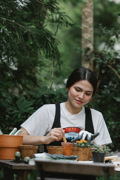 Busy woman in glove with spade planting seedling in peat pot while sitting at table against green plants in garden